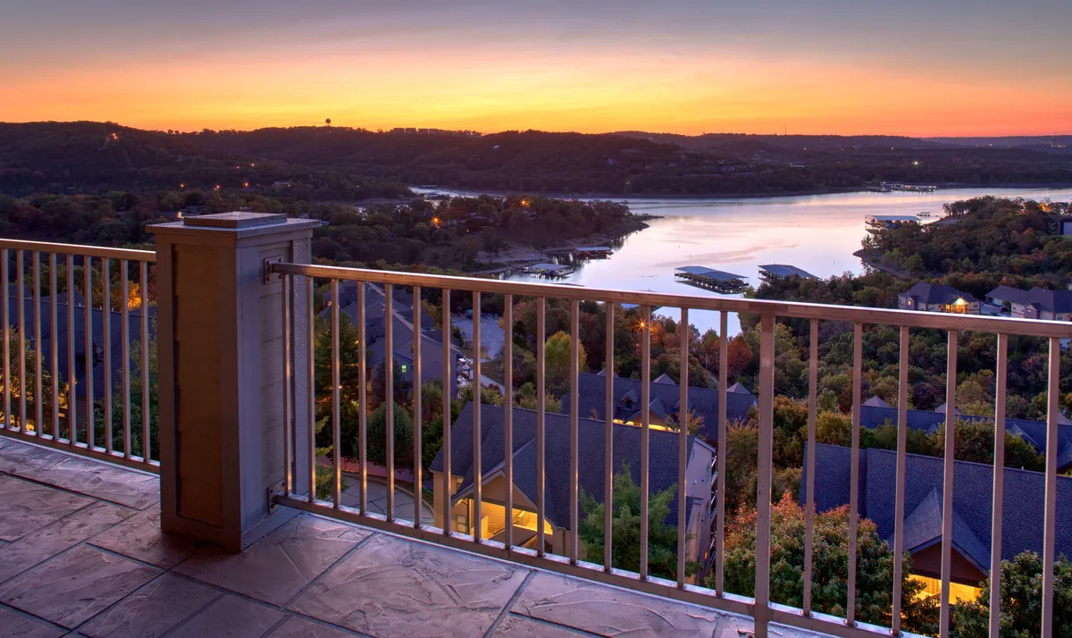 A serene balcony scene with a clear view of the lake and distant hills at sunset. The balcony railing frames the vista, inviting viewers to imagine relaxing or socializing as the sky changes color. The calm mood and open-air setting evoke the exclusivity and tranquility referenced in the content.