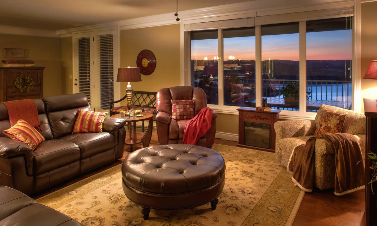 A spacious, open-concept living room featuring large windows that frame a stunning lake sunset. Comfortable leather sofas, a stylish armchair, and a round ottoman are arranged around a decorative rug. The inviting electric fireplace anchors the space, creating a cozy atmosphere ideal for relaxing, socializing, or movie nights. The design seamlessly blends warmth and style while drawing the eye toward the panoramic lake views.