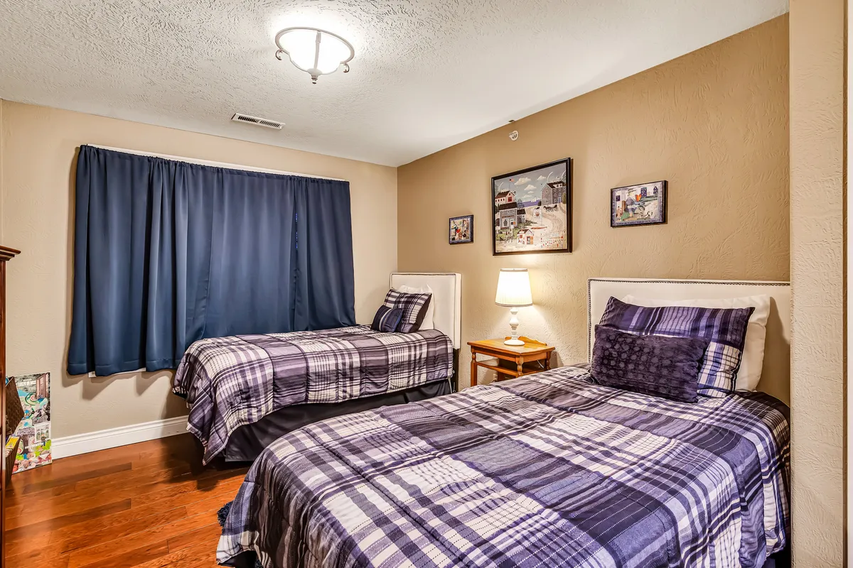 A cheerful and comfortable bedroom with twin beds, a nightstand and lamp, blue curtains, and framed artwork. The room feels welcoming and is ideally suited for children or friends seeking their own space.