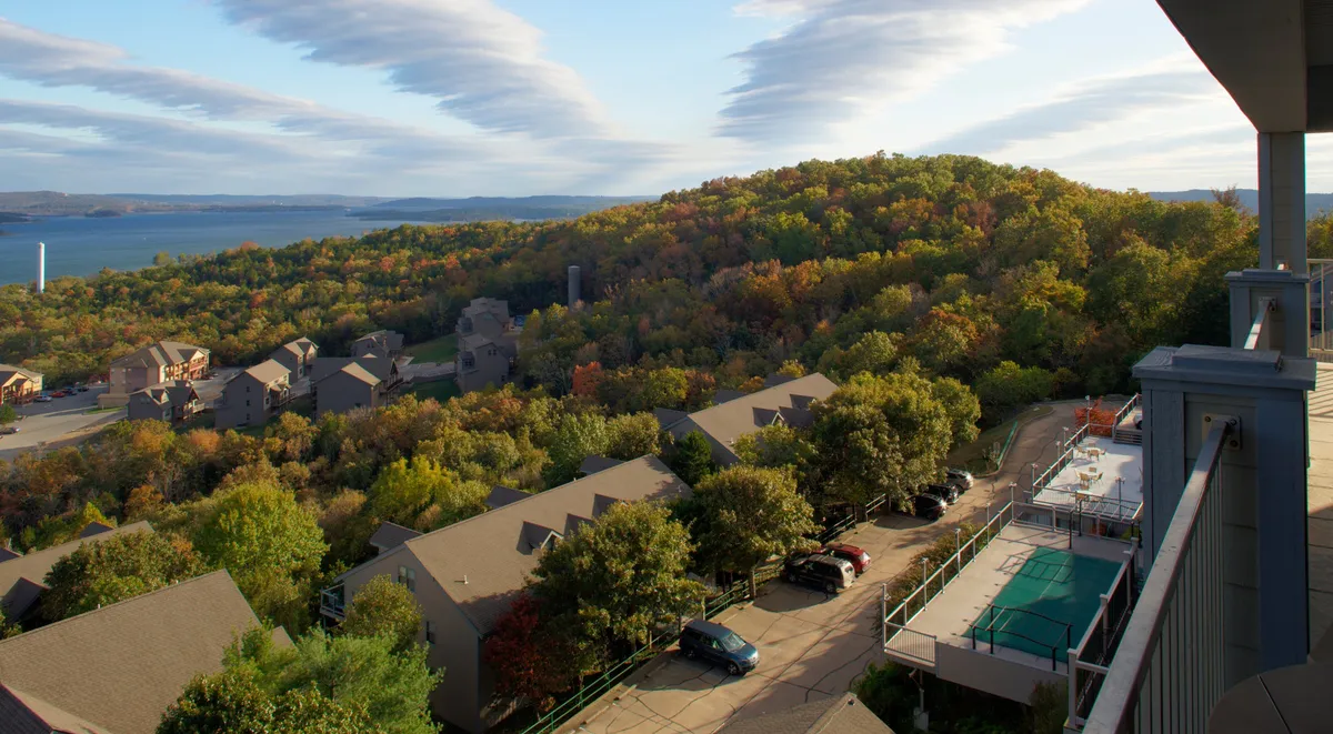 A wide view of the resort community, featuring colorful autumn trees, the shimmering lake, residential buildings, and the swimming pool area. The image illustrates both the natural beauty and the shared amenities available to guests—such as the pool and walking trails—within the setting of the larger community.
