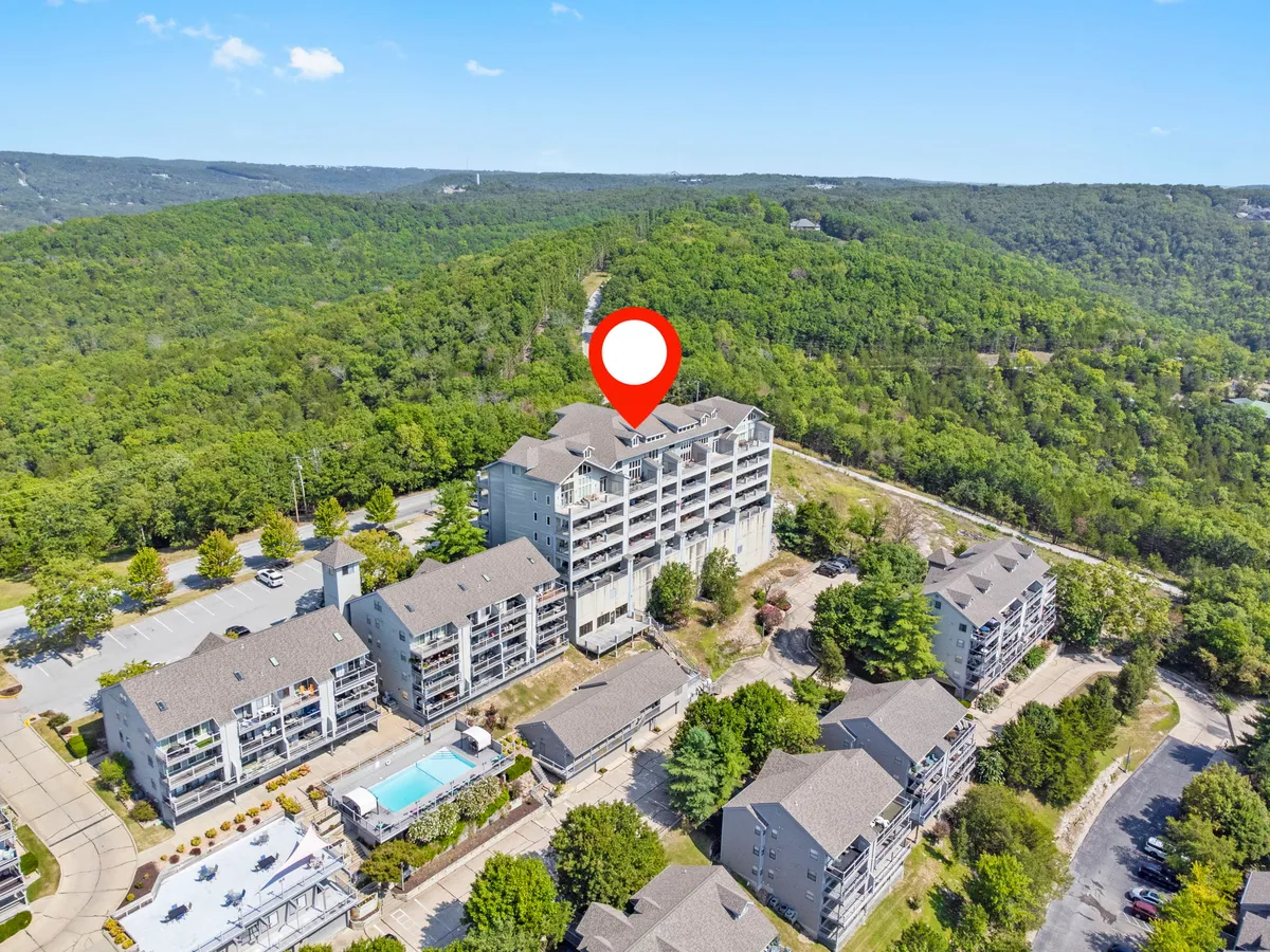 An expansive view of the condo’s multi-story building, lush greenery, the outdoor swimming pool, a parking area, and a road that leads to the property. The image conveys both the community feel and easy access to walking trails and amenities.