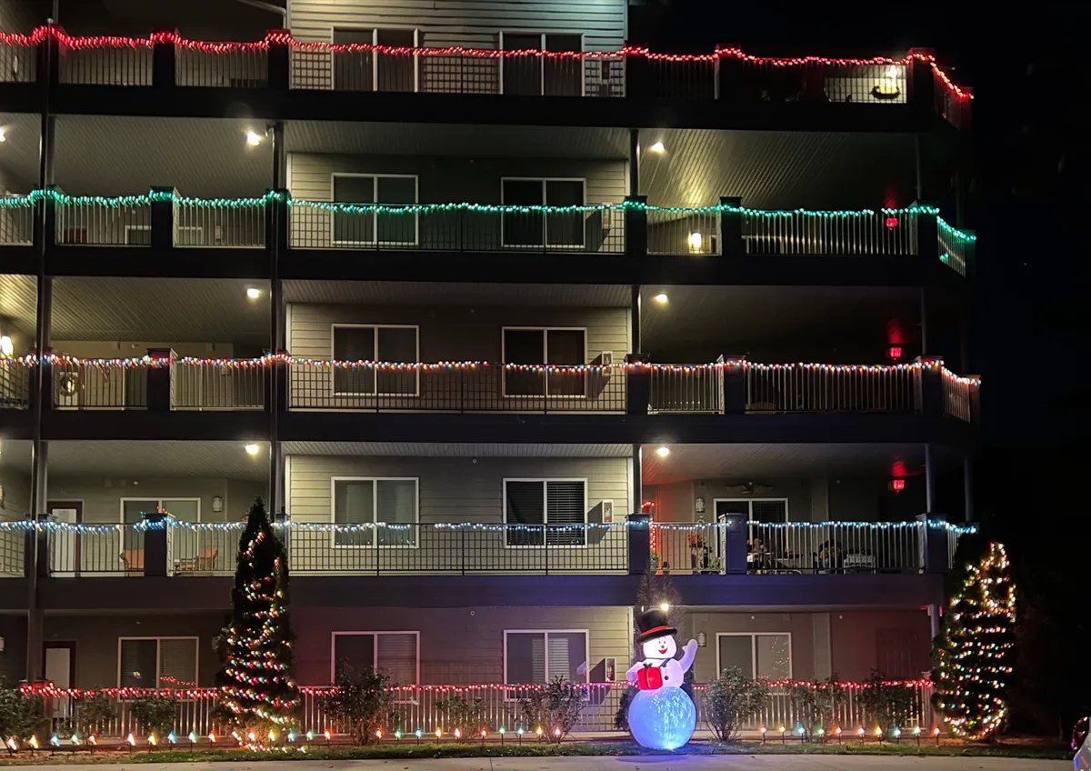 A festive exterior shot featuring decorative holiday lights strung across balconies, a cheerful snowman decoration, and a Christmas tree. The atmosphere is lively and inviting, evoking the spirit of community holiday celebrations.